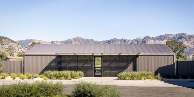 A single-storey contemporary building with a long, simple rectangular form and low-pitched metal gabled roof. The dark vertically ribbed facade is screened by horizontal slatted elements around the central glazed entry, with landscaped grasses and paving in the foreground. Rugged mountains form the distant background.