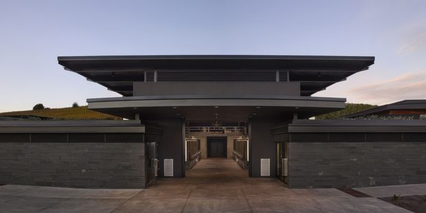 A symmetrical contemporary residential entrance viewed from outside at dusk. Dark masonry boundary walls and broad horizontal roof planes frame a recessed pedestrian entry, with linear louvred detailing and low planted roof areas visible on the adjoining structures. The composition emphasizes strong horizontal lines, deep overhangs, and a dark monochromatic material palette.