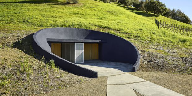 A partially earth-sheltered contemporary structure embedded into a grassy hillside. The building has a smooth dark curved concrete or rendered shell forming a recessed forecourt, with a flat roof, timber-clad entry doors, a large glazed opening, and a concrete-paved path leading to the entrance. Vineyard-like rows and landscaped rural terrain surround the structure.
