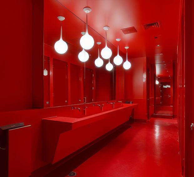 A dramatic monochromatic red restroom interior with a long angular built-in vanity, multiple integrated basins and faucets, wall-to-wall mirrors, and a row of sculptural white pendant lights. Red doors, walls, ceiling, floor, and joinery create a bold, immersive commercial washroom design, with a corridor extending toward the rear.