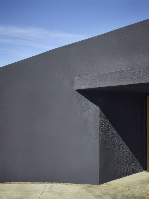 Minimalist architectural exterior showing a dark charcoal rendered facade with sharp geometric planes and a deeply recessed entry or opening. A narrow warm-toned door is partly visible at the right, contrasting with the textured dark surface. The low, angular roofline and strong shadow create a stark, sculptural composition against a clear blue sky.