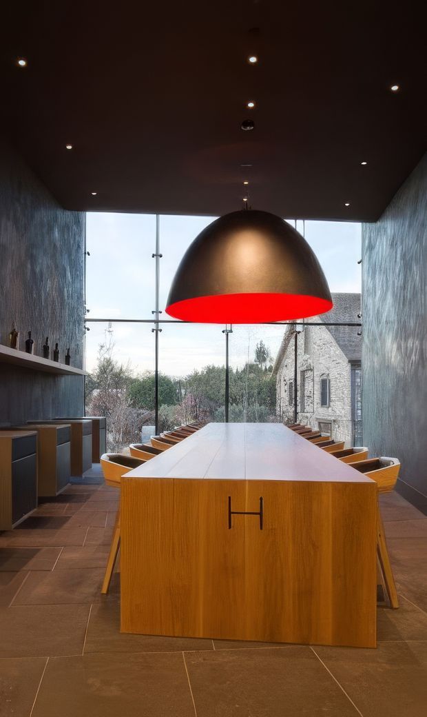 A dramatic contemporary dining room with a long central timber dining table and integrated storage end, surrounded by sculptural wooden chairs. A large metallic dome pendant with a vivid red underside hangs above the table. Dark textured walls and ceiling create an intimate, moody atmosphere, while full-height glazing at the far end overlooks trees and a stone building. Large-format brown stone or porcelain floor tiles complete the space.