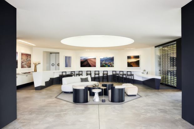 A spacious, contemporary open-plan interior with a lounge and bar or dining area. The room has a polished concrete floor, white walls and ceiling, dark navy or black architectural wall panels, a large oval recessed ceiling light, and extensive built-in bar cabinetry with stools along the rear wall. A curved arrangement of white and dark cylindrical lounge furniture sits on a geometric rug, with framed artwork and a patterned glass or screen wall adding visual interest.