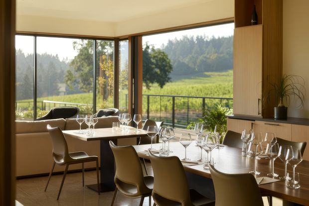 A warm, contemporary open-plan dining and living space with floor-to-ceiling glazing overlooking a green rural landscape. The room features a large formally set dining table, sculptural upholstered dining chairs, a secondary dining table, a neutral sofa, dark timber or stone-look built-in cabinetry, and extensive timber-framed windows opening toward a balcony or terrace.