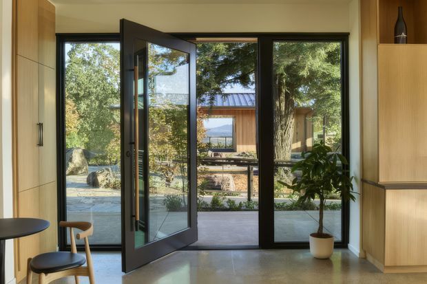 A refined contemporary entryway or transitional interior looking through dark-framed glazed doors toward a landscaped terrace and a separate timber-clad structure. The space has light timber built-in cabinetry on both sides, a polished pale floor, warm off-white walls, and a potted indoor tree. The open pivot or hinged glazed door creates a strong connection to the outdoor setting and distant landscape.