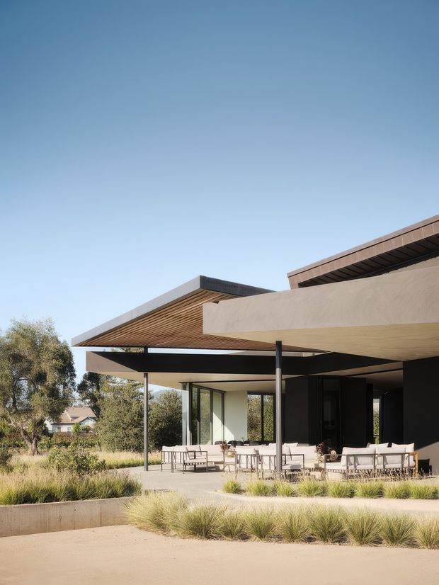 Exterior view of a contemporary low-profile residence with projecting flat roof planes, deep timber-lined eaves, slim dark structural posts, and extensive floor-to-ceiling glazing. A furnished outdoor terrace and landscaped drought-tolerant garden occupy the foreground, with mature trees and a neighbouring house beyond.