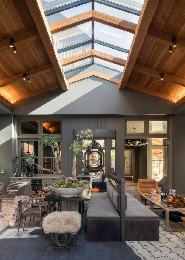 A sophisticated open-plan interior with a dramatic timber-lined pitched ceiling and large central skylight or clerestory glazing. The room combines a dining area, lounge seating, dark built-in joinery, grey wall panelling, warm timber beams, patterned rugs, and sculptural decorative furnishings. Abundant daylight contrasts with warm recessed and pendant lighting, creating a refined, layered atmosphere.