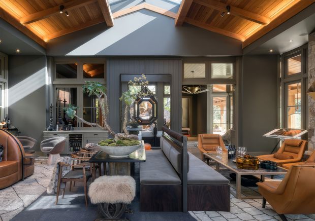 A sophisticated open-plan living room with dark charcoal walls, extensive warm timber ceiling lining and exposed structural beams. The room features a central skylight, high pitched ceiling, large glazed openings, a dark built-in sectional sofa, leather lounge chairs, a glass coffee table, indoor plants, and a decorative mirrored cabinet. Strong daylight creates dramatic geometric shadows across the walls and furnishings.