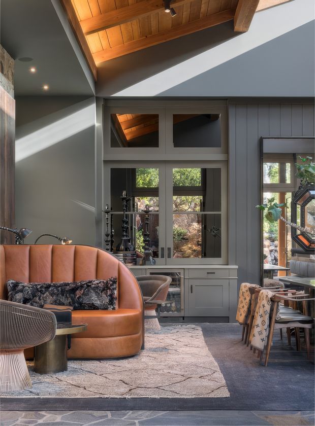 A stylish open-plan living and dining area with a dramatic pitched ceiling featuring exposed timber lining and beams. The room has dark grey-blue wall panelling, grey cabinetry beneath a multi-pane window, a rust-orange curved leather sofa, patterned cushions, sculptural lounge chairs, a dining table, and a large neutral rug over dark flooring. Strong sunlight enters through the glazing, creating bold diagonal shadows and highlighting the warm timber ceiling. A partially visible adjoining dining area and exterior garden are seen through the windows.