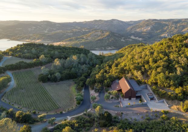 Aerial view of a rural mountain property surrounded by wooded hills, cultivated rows, winding roads and a lake or reservoir in the background. A compact building complex with a pitched roof sits on a cleared site amid the landscape.