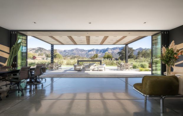A spacious contemporary open-plan interior with polished concrete flooring and dark walls opening through fully retracted glazed doors to an expansive outdoor terrace. A home office or work area with multiple office chairs and desks occupies the left side, while a lounge chair and plants sit to the right. The covered terrace features outdoor seating and dining, with dramatic mountain scenery visible beyond. The ceiling combines a smooth interior plane with a timber-lined covered outdoor soffit, creating a strong indoor-outdoor connection.
