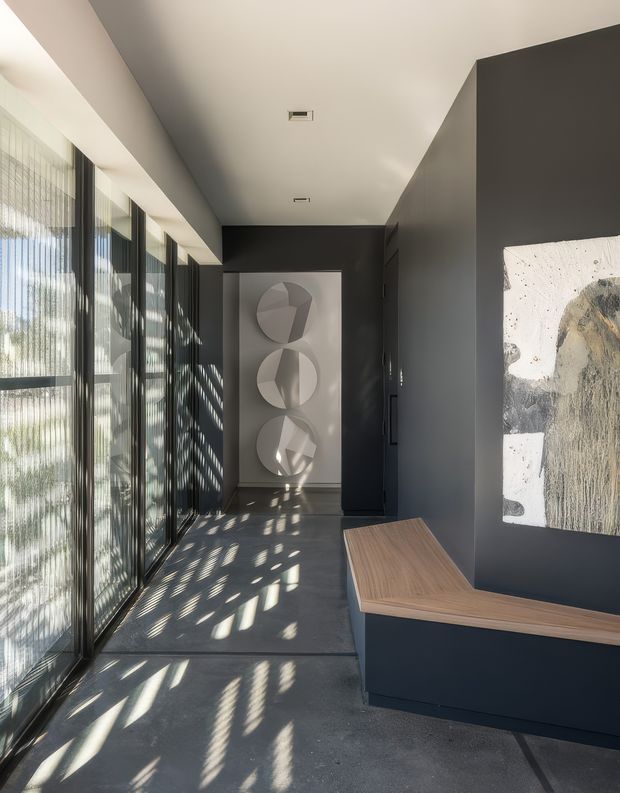 A refined contemporary interior corridor or entry gallery with full-height glazed windows and vertical sheer blinds along one side, casting patterned sunlight across a dark polished floor. Charcoal walls and ceiling create a dramatic backdrop for abstract artwork and sculptural circular wall pieces. A built-in dark joinery element with a warm timber benchtop projects into the foreground, likely functioning as a console or reception-style counter. The space is minimalist, high-contrast, and gallery-like.