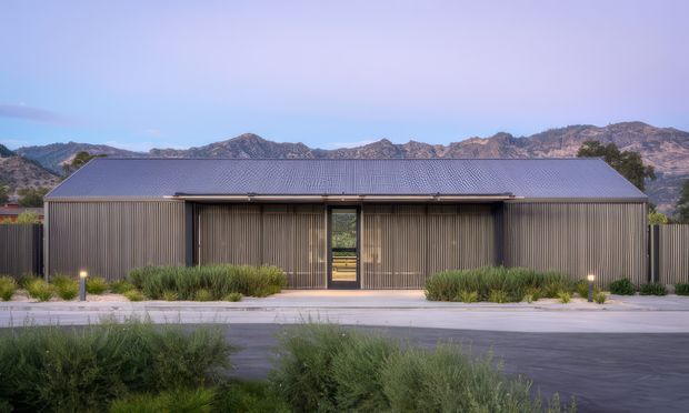 A single-storey contemporary building viewed from the landscaped street frontage. The long rectangular structure has a dark corrugated metal gable roof, vertically ribbed dark metal cladding, and a recessed central glazed entry framed by deep horizontal eaves. Low ornamental planting and pathway lighting line the front, with rugged mountains visible behind the building.