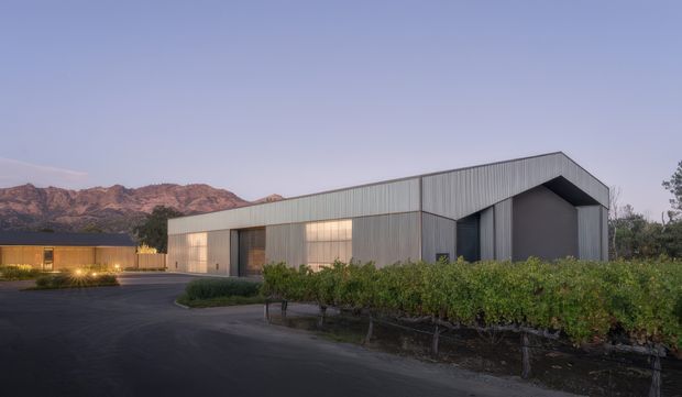 A low, modern metal-clad building viewed at dusk, with a sharply angled gabled roof and large glazed openings. It sits beside a landscaped vineyard or planted rows, with rugged mountains visible in the background and a paved forecourt in the foreground. Warm interior lighting contrasts with the cool blue evening sky.