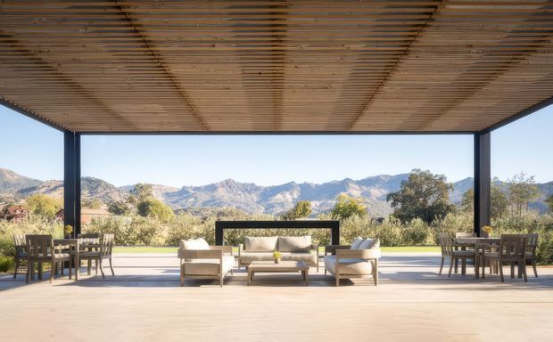 A spacious covered outdoor entertaining terrace with a timber slatted ceiling and dark slender structural posts. Lounge seating and two dining settings are arranged on a light timber or composite deck, opening onto expansive mountain and landscaped views. The design is open-sided, minimalist, and strongly connected to the surrounding landscape.