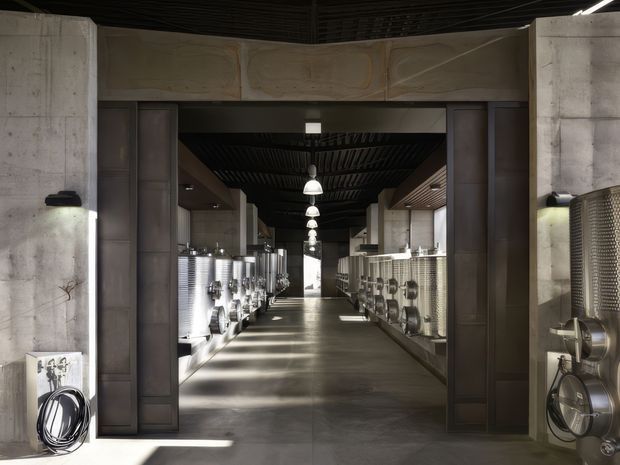 A symmetrical industrial brewery or production facility interior viewed through a large dark metal-framed opening. Stainless-steel brewing tanks and processing equipment line both sides of a long polished concrete corridor, with exposed concrete walls, dark ribbed ceiling panels, suspended pendant lights, and strong linear daylight. The scene has a utilitarian, contemporary-industrial character.