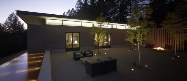 A low-slung modern house viewed from an expansive paved courtyard or terrace at dusk. The dark, minimalist facade has a long horizontal clerestory window band beneath a projecting flat roof, glazed doors opening to the courtyard, and discreet ground lighting. Outdoor lounge furniture sits in the foreground, while a tall timber privacy screen and mature trees frame the site.
