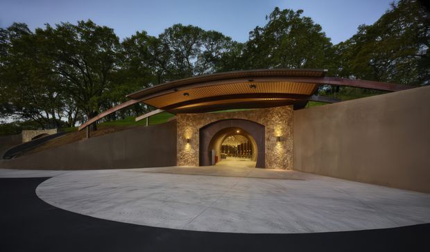 A dramatic exterior entrance set into a landscaped, tree-filled site. A broad curved timber-lined canopy projects over a stone-clad arched passageway, with warm recessed lighting illuminating the entry. Smooth rendered boundary walls and a large pale concrete forecourt contrast with the dark surrounding paving and mature trees.