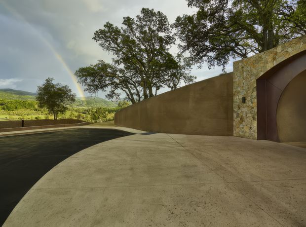 Exterior architectural view of a broad concrete driveway or forecourt curving alongside a tall earthen-toned rendered wall. At the right is a substantial stone-clad entry structure with a dark brown arched opening or gate. Mature trees rise above the wall, while cultivated rural hills and vineyards are visible beyond. The scene has a restrained, earthy, sculptural character under overcast light, with a faint rainbow in the background.