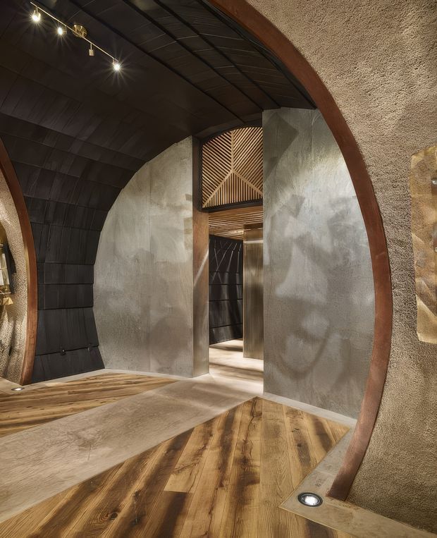 Dramatic interior corridor or entry space with large rounded arch openings, dark ribbed metal ceiling and wall cladding, raw textured plaster or concrete surfaces, warm timber flooring, and sculptural timber slatted detailing visible through the opening. Recessed and track lighting accentuate the highly crafted, cave-like architectural interior.