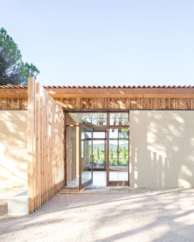 A bright exterior view of a low-rise modern building with a warm timber slatted facade, pale rendered walls, a shallow terracotta tiled roof, and large glazed timber-framed doors opening into a light-filled interior. Gravel paving and strong tree shadows surround the entrance, with garden vegetation visible beyond.