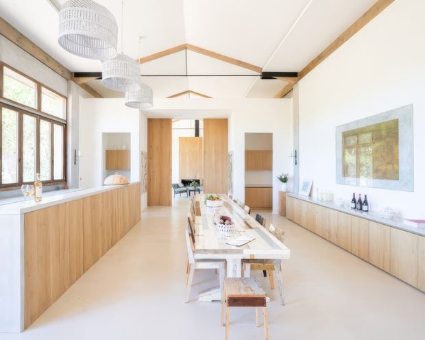 Bright, open-plan kitchen and dining space with extensive light timber cabinetry, white benchtops, a long central dining table, and large timber-framed windows. The room has a high vaulted ceiling with exposed timber beams and several white pendant lights. A minimal, warm contemporary fit-out includes a low built-in sideboard, open shelving, and a large artwork.