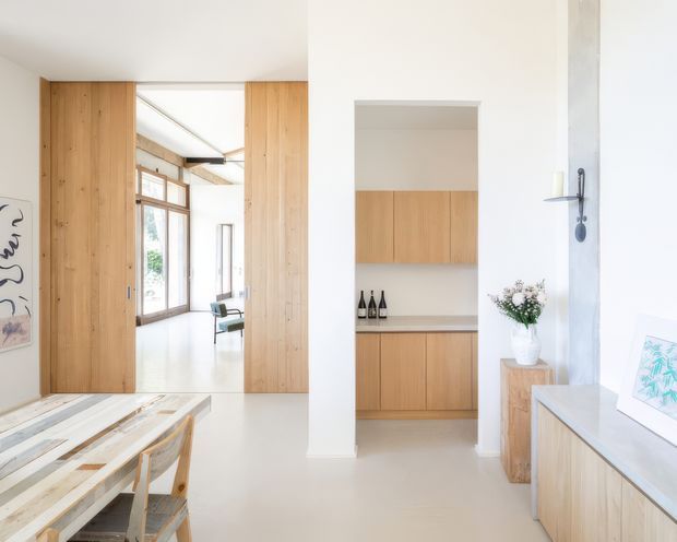 Bright, minimalist interior showing a dining area in the foreground, opening through full-height timber joinery into a glazed living space. A compact built-in timber storage or bar cabinet is recessed into the white wall, with a pale benchtop and open shelving. The room has white walls and ceiling, light flooring, warm natural timber cabinetry, and abundant daylight. A partial artwork and decorative objects are visible, with a large window or glazed opening on the right.