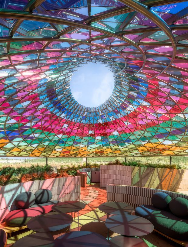 A vibrant covered outdoor lounge or courtyard beneath an elaborate translucent stained-glass-like canopy. The canopy forms a dramatic spiralling oval opening to the sky and is supported by a dense curved metal framework, casting colourful geometric shadows across curved built-in seating, low round tables, and tiled flooring. Pink, red, turquoise, blue, yellow, and green panels create an immersive, kaleidoscopic atmosphere, with planted beds around the perimeter.