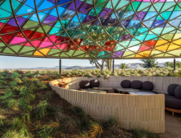 An outdoor covered lounge area with curved built-in seating arranged around a low central table. The space is defined by a striking geometric canopy made from multicoloured translucent glass or panels in turquoise, blue, purple, red, yellow and green, supported by a dark metal framework. Naturalistic planting surrounds the curved pale wall and seating, with a broad landscape visible beyond.
