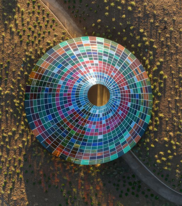 Aerial view of a large circular, radial structure or artwork composed of concentric rings and wedge-shaped panels in vivid blue, teal, turquoise, red, orange, white, and grey. A circular opening occupies the centre, with a timber-toned surface visible below. The structure is set within a landscaped or agricultural site crossed by curved paths.