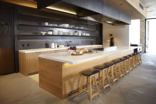 Spacious contemporary kitchen with a large timber-clad island and integrated seating for a long row of stools. The kitchen features a pale stone benchtop, dark open shelving and splashback, extensive stainless-steel rangehood structures, timber ceiling detailing, and large-format tiled flooring. The room is open and bright, with a glimpse of an adjacent area through the opening at right.