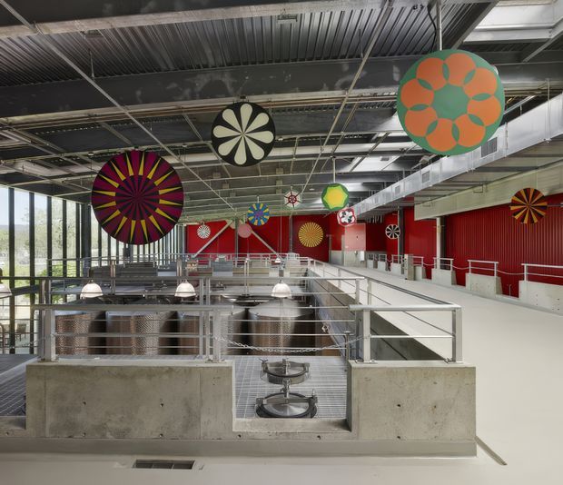 A large industrial-style commercial interior with an open mezzanine or elevated walkway overlooking a lower work or dining area. The space features exposed corrugated metal ceilings, visible services and ductwork, red corrugated wall cladding, polished metal railings, concrete parapets, gridded metal flooring, and extensive floor-to-ceiling glazing. Large colourful circular graphic panels and pendant lights add playful visual accents.