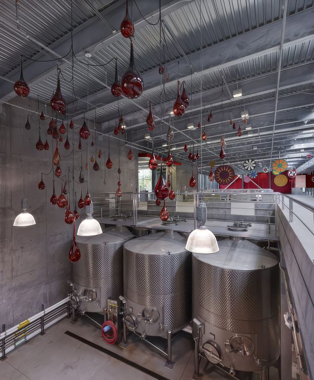 Interior view of an industrial brewing or production facility, showing several large stainless-steel cylindrical tanks in the foreground, exposed services and corrugated metal ceiling overhead, suspended pendant lights, and numerous red glass teardrop ornaments hanging throughout the space. A colourful mural or decorative wall installation is visible in the background. The space has a utilitarian, adaptive industrial character.