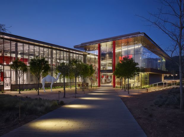 Evening exterior view of a contemporary multi-storey glazed building complex. The architecture features extensive floor-to-ceiling curtain glazing, projecting flat roof planes, exposed structural elements, illuminated interiors, and vivid red vertical columns. A landscaped pedestrian approach with paving, young trees, lighting, and a white sculptural horse leads toward the entrance.