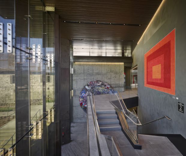A contemporary interior stairway and circulation space with exposed concrete walls, dark linear timber ceiling, large glazed doors, stainless-steel handrails, and a bold red geometric artwork. The stair descends beside a sculptural patterned installation, with subdued integrated lighting and a refined commercial or institutional character.