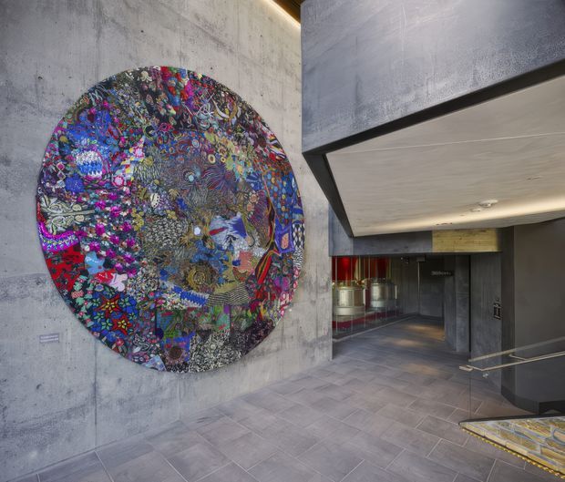 A contemporary interior lobby or gallery-like circulation space with raw grey concrete walls, a large colourful circular artwork mounted on the feature wall, dark structural framing, recessed warm lighting, and grey tiled flooring. A stair or ramp descends toward a darker lower-level area, with glimpses of a bar or service zone beyond.