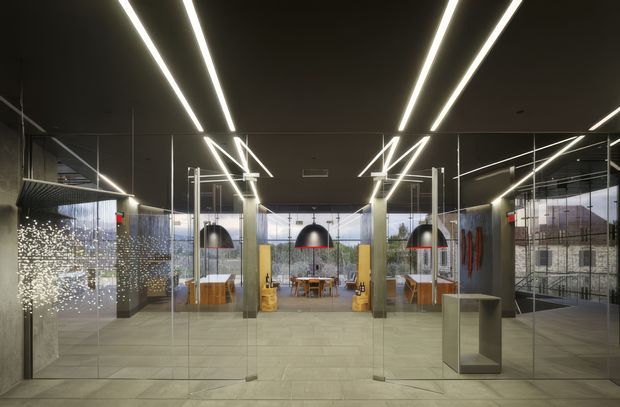 A contemporary commercial interior, likely an office reception or collaborative workspace, viewed through extensive clear glass partitions. The space features a dark ceiling with long linear LED lighting, polished concrete or large-format grey floor tiles, exposed structural columns, suspended black pendant lights, timber meeting tables and cabinetry, and glazed office rooms beyond. Reflections and a bright decorative light installation are visible in the glazing.