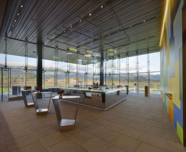 A spacious, contemporary commercial interior with full-height glass walls overlooking a landscaped valley and hills. The room features a reflective slatted ceiling with recessed spotlights, large square tiled flooring, a central angular service or display counter, sculptural geometric tables and chairs, and a colourful abstract wall installation along the right side.