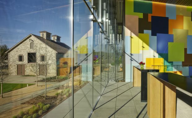 A contemporary commercial interior with full-height glazed walls, polished concrete flooring, and a bold geometric feature wall composed of colourful rectangular panels. Timber-fronted counters or built-in joinery occupy the foreground. Through the glazing, a traditional stone-clad building and landscaped grounds are visible, creating a strong contrast between historic exterior architecture and modern interior fit-out.