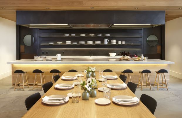 A refined open-plan kitchen and dining space centered on a long light-wood island with integrated seating. The kitchen features dark built-in cabinetry and open display shelving, a pale stone benchtop, stainless-steel appliances and hardware, and warm under-cabinet lighting. In the foreground, a large timber dining table is fully set with plates, glasses, cutlery, and floral arrangements. The ceiling is clad in warm timber with recessed spotlights, creating a polished, inviting atmosphere.