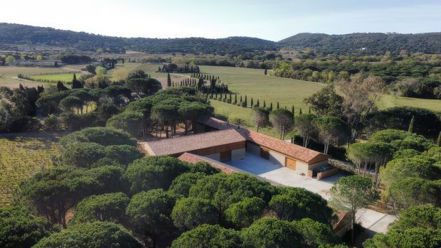 Aerial view of a low-profile residential property set within a broad rural landscape. The home is arranged around a central rectangular courtyard or pool-like open space, with warm terracotta tiled roofs, earthy walls, and extensive mature greenery. Vineyards or cultivated fields, rows of slender trees, and wooded hills surround the property.