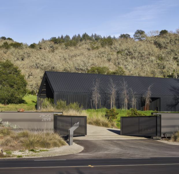 A contemporary dark-clad building viewed from the road, set behind landscaped planting and automated black gates. The building has a long, steeply pitched gable roof with standing-seam metal roofing and dark vertical cladding. A hillside of mature vegetation rises behind it, and a rendered boundary wall displays the address numbers 2900 and 2898.