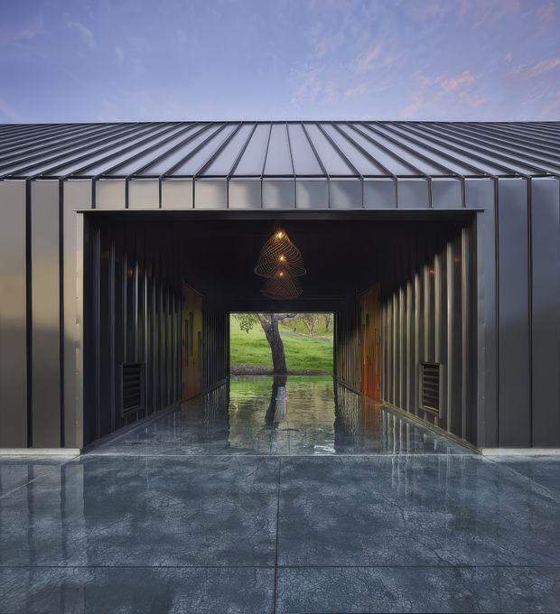 Front-facing view of a dark, contemporary metal-clad building with a broad pitched standing-seam metal roof. A deep central passage or breezeway leads through the structure, with reflective polished concrete paving, vertical ribbed wall surfaces, warm timber doors, integrated wall vents, and a sculptural pendant light. The passage frames a tree and green landscape beyond, while the wet or highly reflective floor creates a strong symmetrical composition.