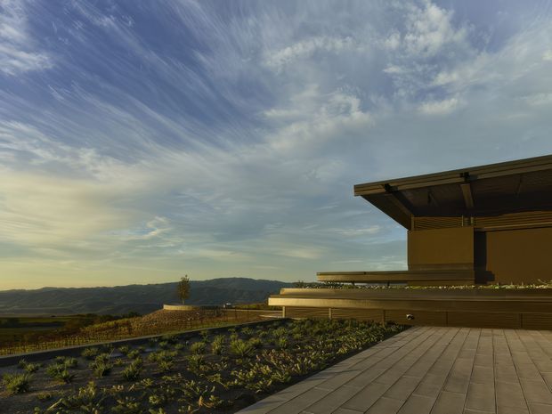 A low-slung contemporary building viewed from a broad paved terrace, with deep projecting roof eaves, horizontal architectural lines, and a planted roof or landscape bed in the foreground. The structure overlooks a rolling rural or vineyard-like landscape beneath a dramatic cloudy sky.