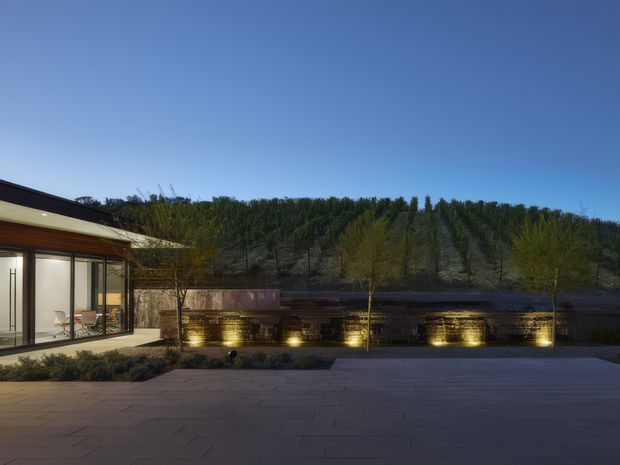 A contemporary single-storey residential exterior at dusk, viewed across a broad paved terrace or courtyard. The building has extensive floor-to-ceiling glazing, dark roof and fascia lines, warm timber-toned soffits, and a low horizontal stone or masonry garden wall illuminated with recessed uplights. A wooded hillside forms the background under a clear blue sky.