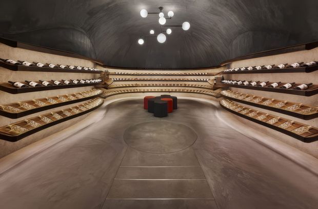 A dramatic, symmetrical wine cellar or tasting room with curved dark plaster walls and ceiling, extensive tiered timber wine racks lining both sides and the rear wall, and a central cluster of low sculptural stools or tables. Warm integrated lighting highlights the wine displays, while a contemporary cluster pendant hangs above.
