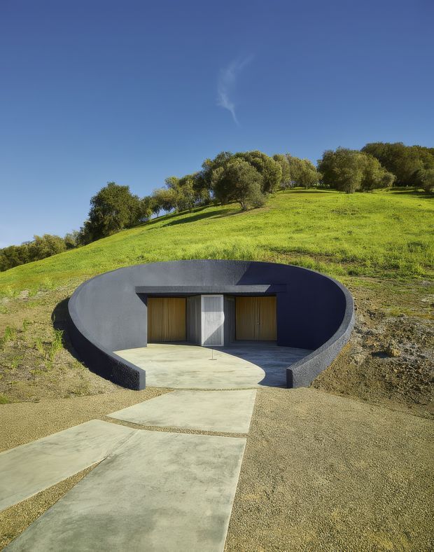 A partially earth-sheltered, sculptural building embedded into a grassy hillside. The dark curved exterior wall forms a recessed semicircular entrance court, with a central glazed door flanked by warm timber vertical screening and a broad concrete pathway leading toward the entry. Rolling grassland and mature trees rise behind the structure under a clear blue sky.