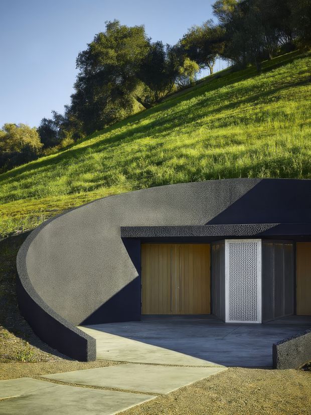 A partially earth-sheltered modern building embedded into a steep grassy hillside. The facade features a dramatic curved dark grey concrete or textured rendered wall, a flat dark roof, a large warm timber garage or entry door, and a patterned metal pedestrian door. Concrete paving leads to the recessed entrance, with mature trees and landscaped grass above.