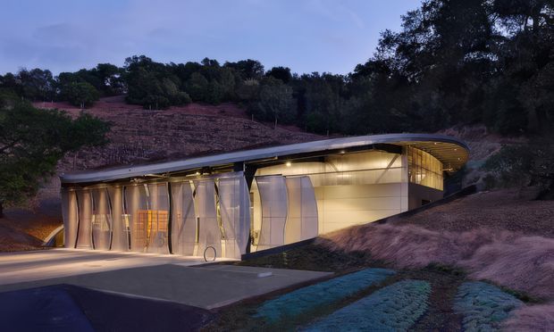 Twilight exterior view of a low, sculptural modern building integrated into a sloping, wooded landscape. The structure has a gently curved roofline, extensive warm-lit glazing, pale metallic wall panels, and a paved forecourt with landscaped planting in the foreground.