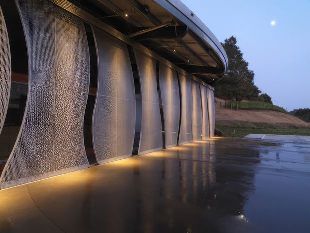 Dusk exterior view of a large contemporary curved building facade. The building features a sweeping roof canopy, exposed dark structural framing, tall curved translucent or perforated metal facade panels, and warm linear lighting at the base. A wet paved forecourt reflects the facade and lighting, with landscaped earth and vegetation beyond.